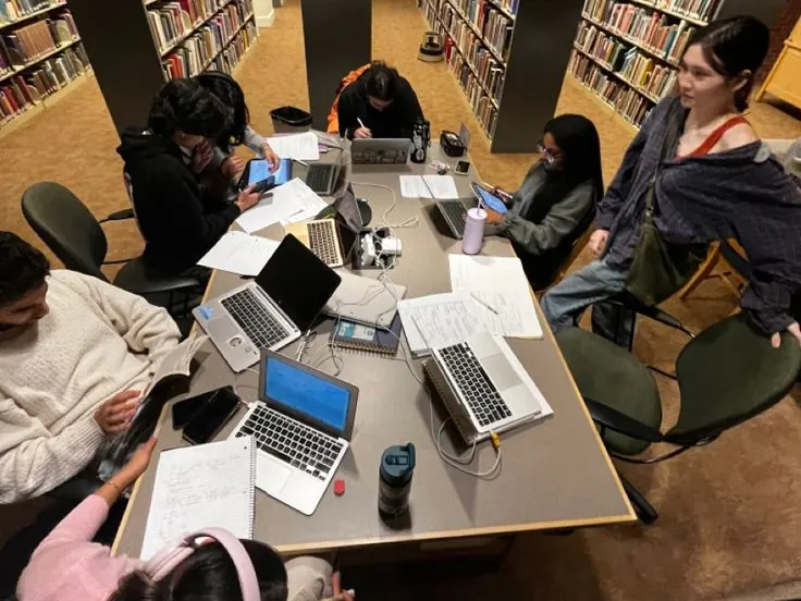 Students working around a table of laptops and papers between library shelves