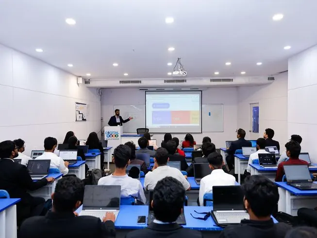 Students at desks with laptops while a lecturer presents at the front of a classroom